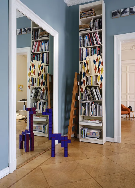 Hohe, gefüllte Bücherregale in blauem Altbauzimmer, davor violette Holzfiguren auf Fischgrätparkett