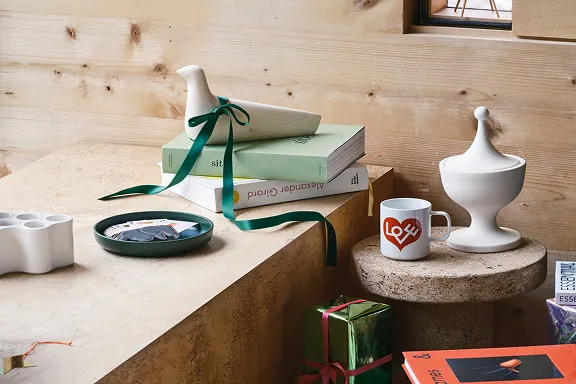 Nahaufnahme: weiße Vogelfigur mit grüner Schleife auf Büchern, Herz-Tasse und weiße Deckeldose auf Korkhocker